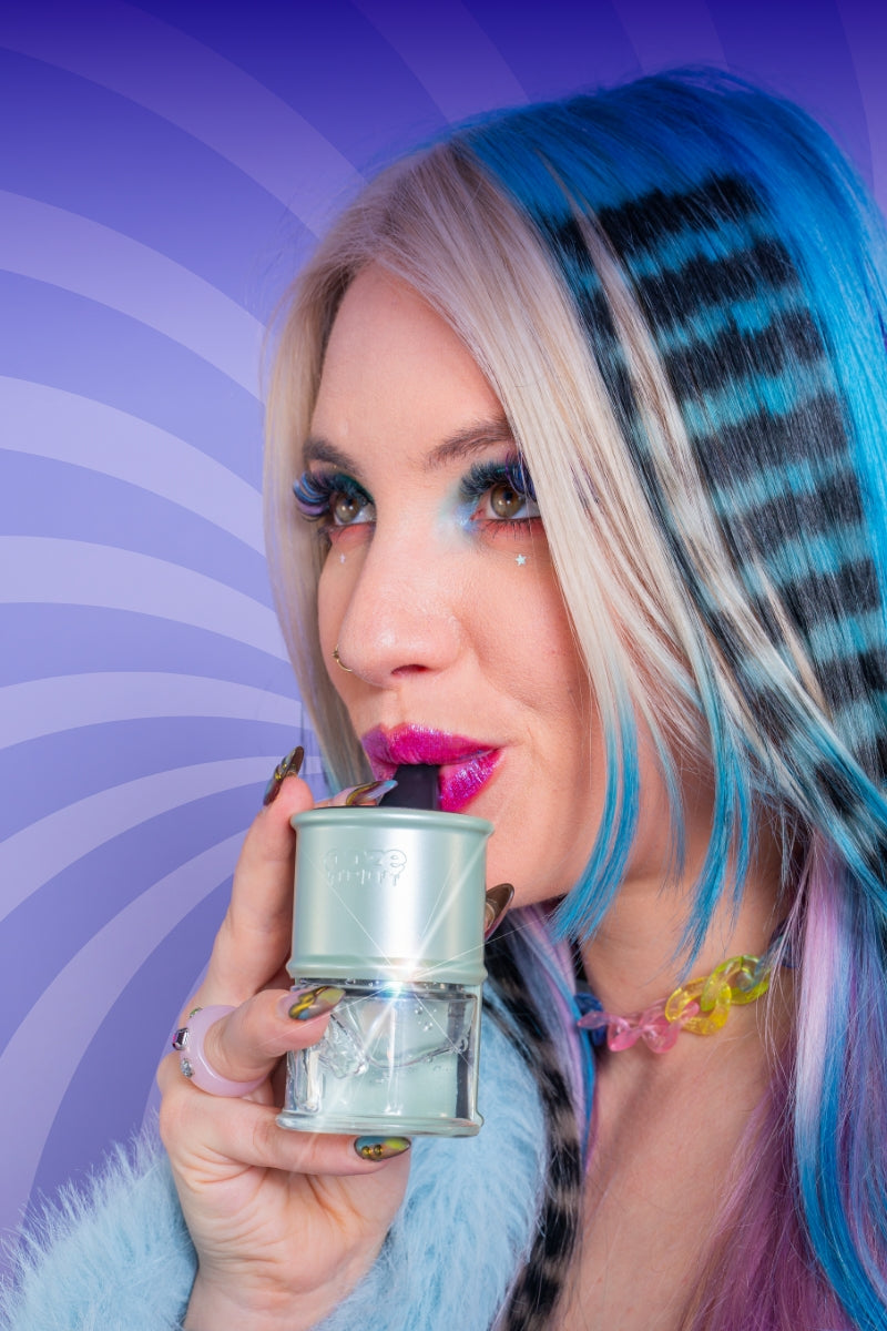 A close-up lifestyle shot of a woman with blue, black, and blonde striped hair taking a dab from a sage green Ooze Electro Barrel e-rig. She is wearing a light blue faux fur collar and a colorful beaded necklace, set against a purple and lavender wavy background.