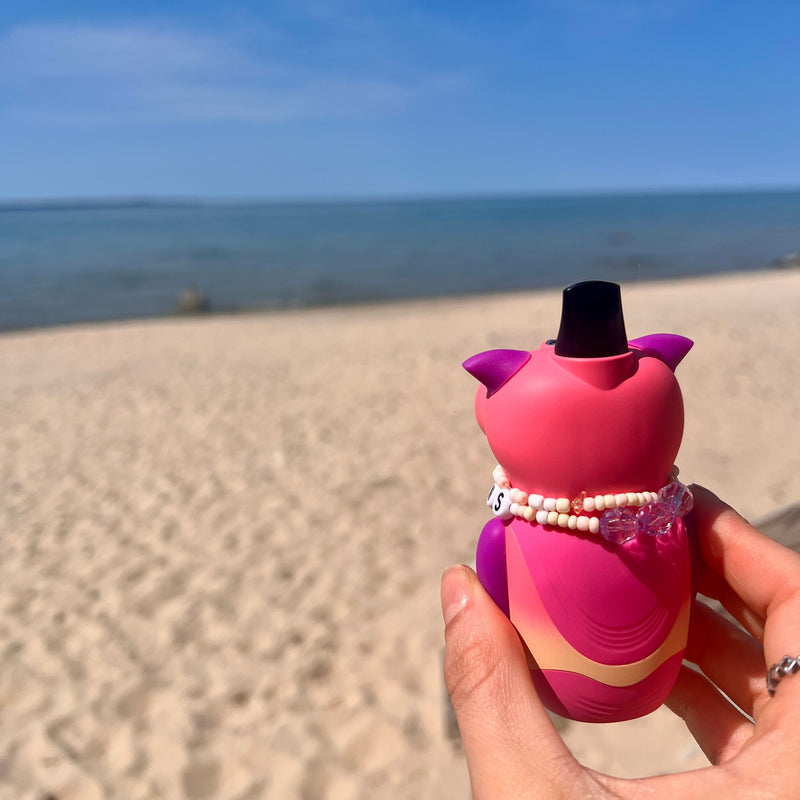 Pink and purple owl shaped device with a black cap held on a sandy beach with ocean and blue sky in the background.