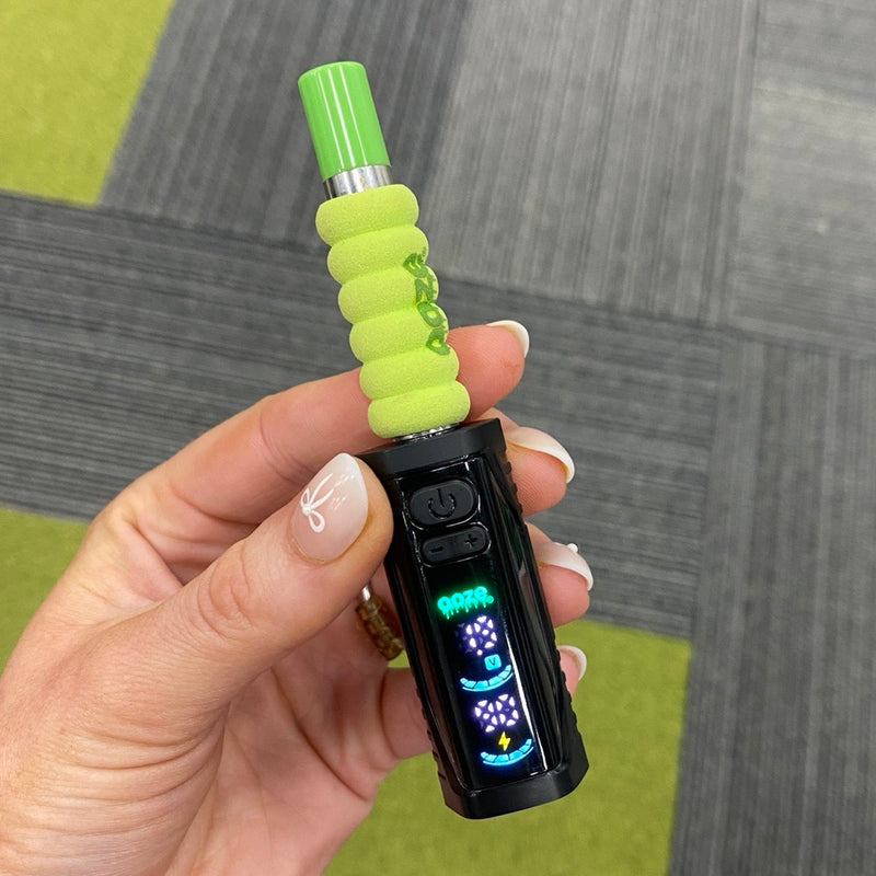 Close up of a person holding the black Ooze Runt Mini Battery with the screen turned on and a cartridge with a green tip and foam cover attached