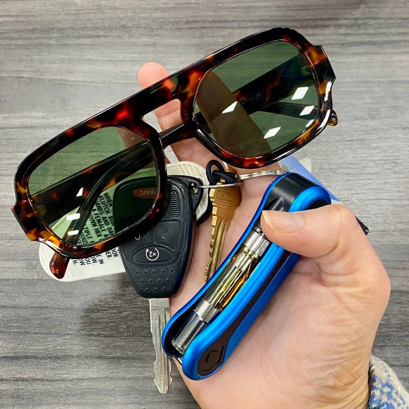 Hand holding sunglasses with a keychain, blue Hinge flick battery, and car keys on a wooden surface