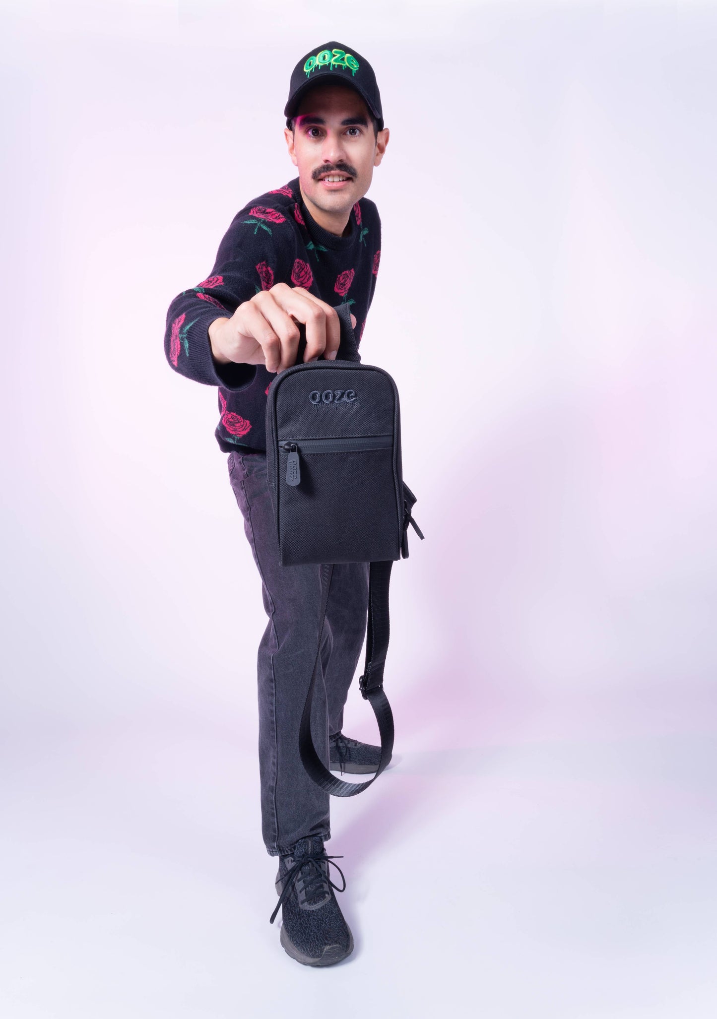 A guy is holding up the black Ooze crossbody bag to the camera. He's wearing an Ooze hat, rose sweater and black jeans