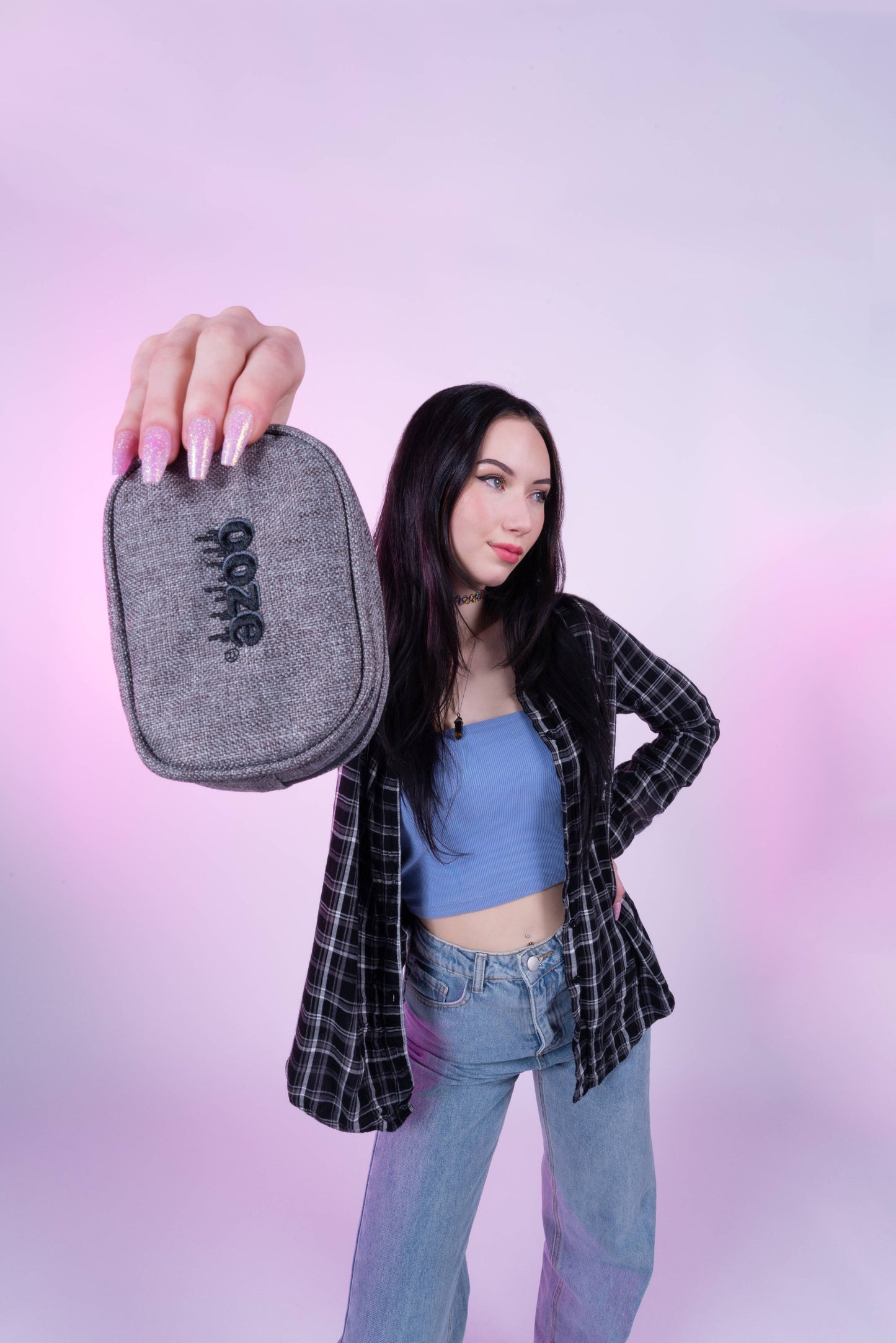 A girl in jeans and a flannel is holding out the smoke grey Ooze wristlet