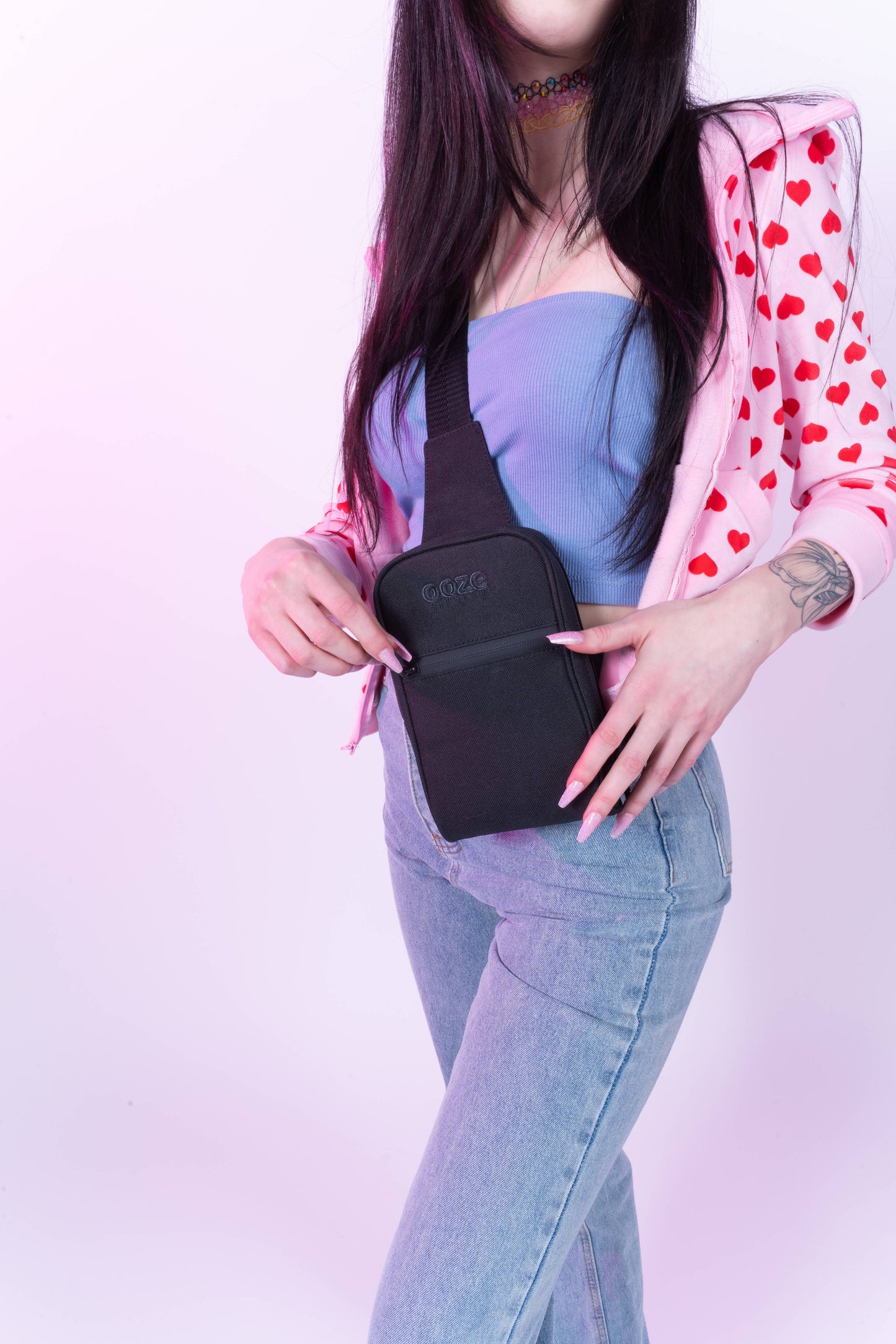 A girl in jeans and a heart zip up hoodie is wearing the black Ooze crossbody bag and about to unzip the front pocket