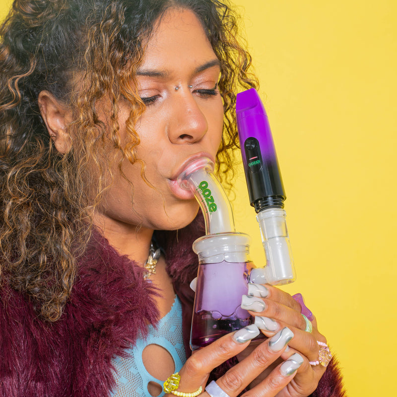 Woman using a purple Booster attached to a purple Glyco freeze bong against a yellow background