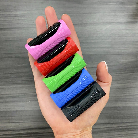 A hand holding five small rectangular Ooze Runt Mini Batteries in different colors: pink, black, red, green, and blue, arranged vertically against a gray wood-textured background.