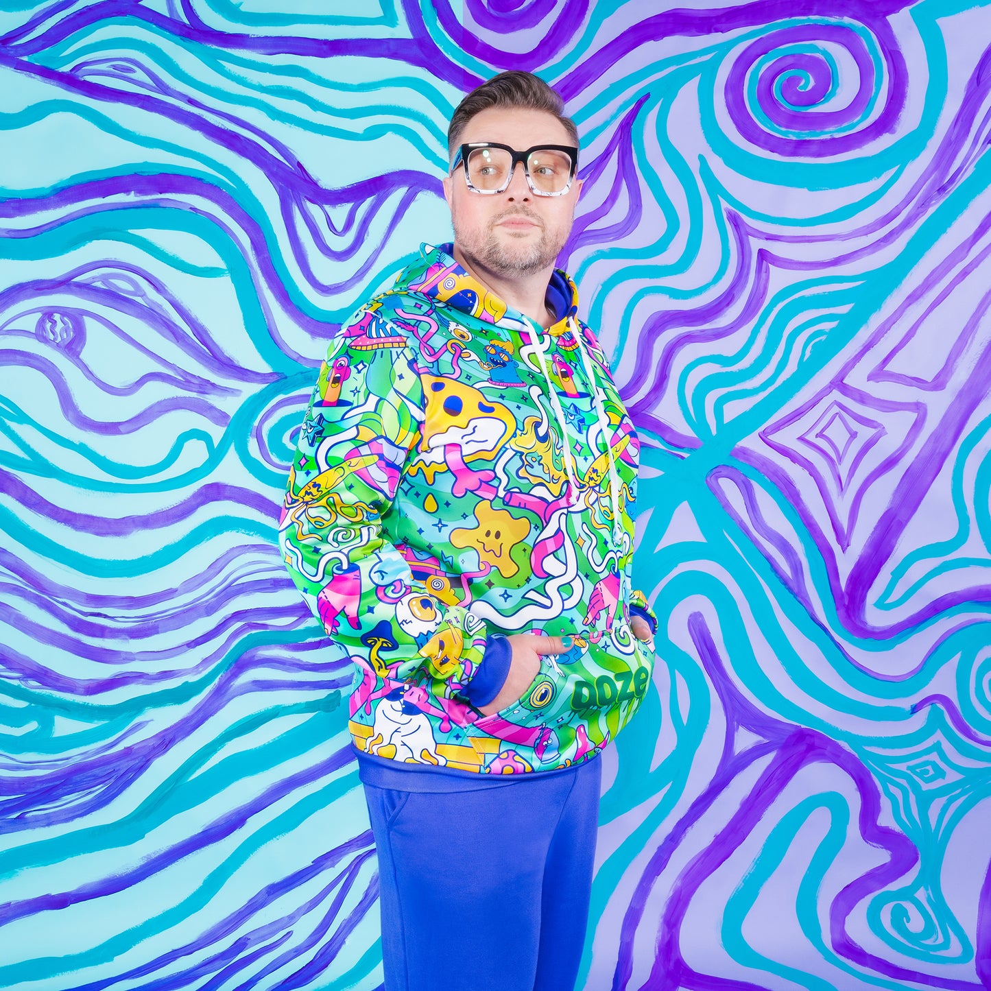 A white guy with chunky square glasses is wearing an Ooze Chroma hoodie. He's looking off in the distance and has his hands in the hoodie pocket.