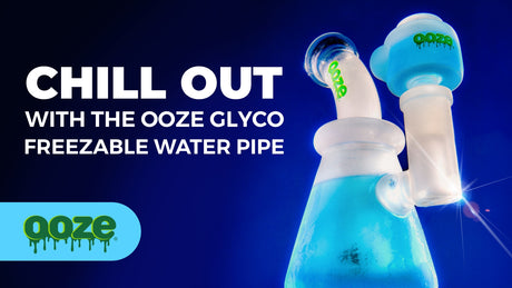 A female hand holds the green Ooze Glyco freezable water pipe. The bong and bowl are frozen, and held up on a deck overlooking some woods, a bench and Lake Michigan.