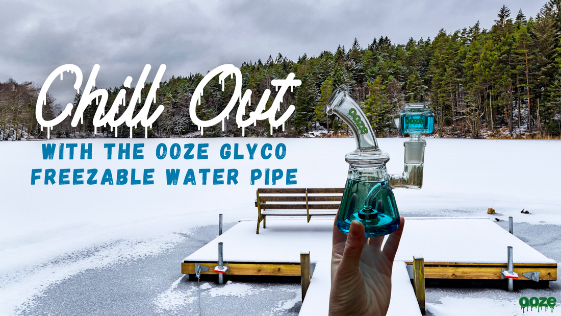 A female hand holds the green Ooze Glyco freezable water pipe. The bong and bowl are frozen, and held up on a deck overlooking some woods, a bench and Lake Michigan.