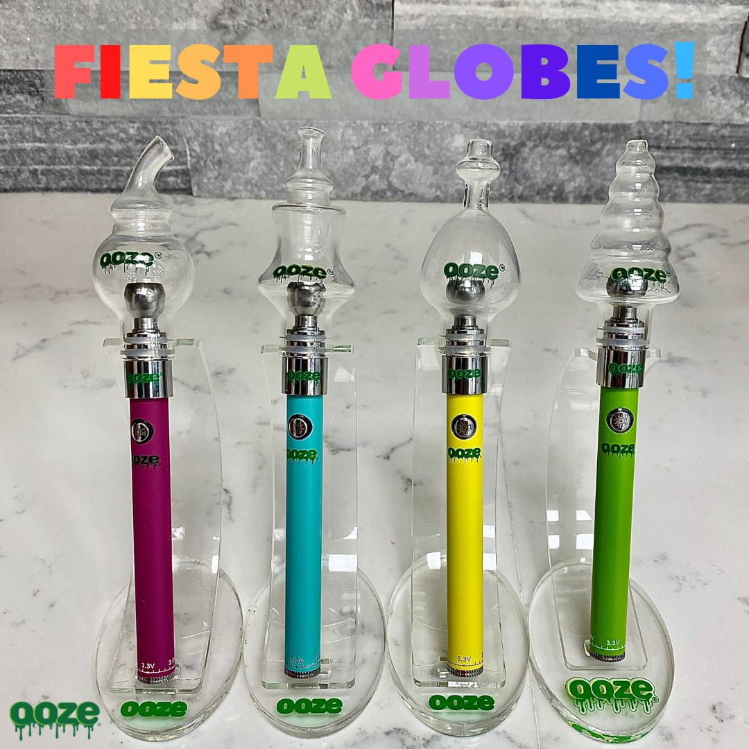 The four new Ooze Glass Globe designs are displayed in a line on a white granite counter. From left to right they are Pink with Swoop Globe, Teal with Genie Globe, Yellow with Cloud Globe, and Green with Festive Globe. All are standing in Showcase Stands.