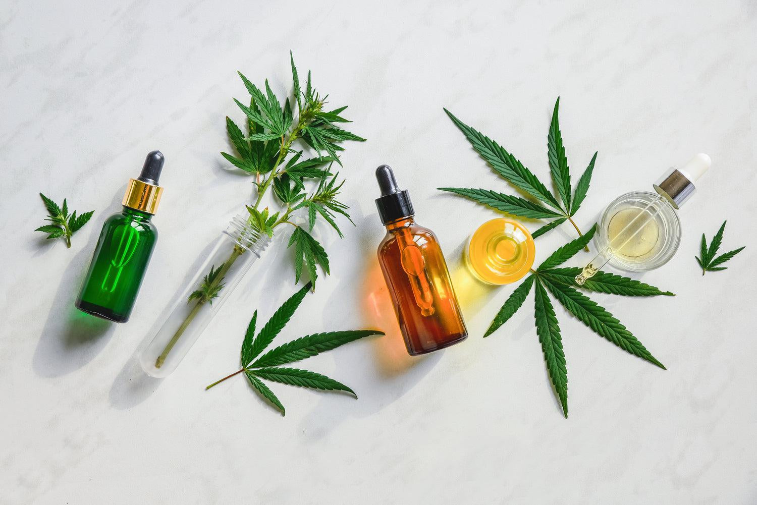 Green, amber, and clear dropper bottles are laying on a white marble background with marijuana leaves placed around them.