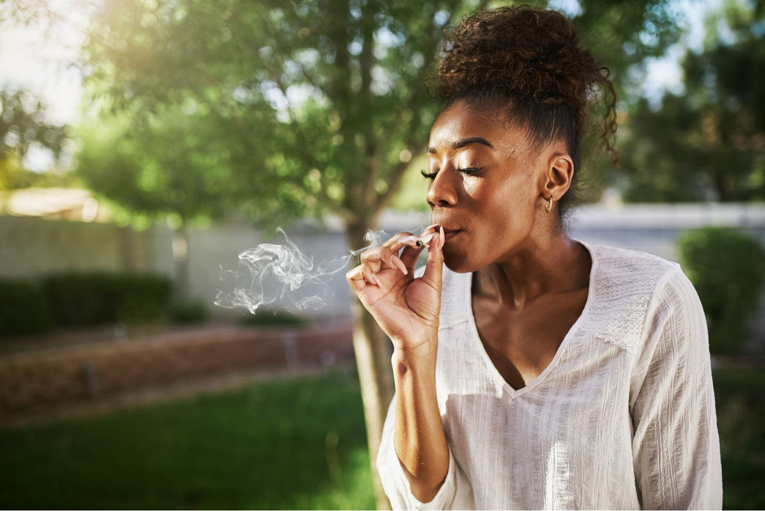 A happy young black woman is smoking a joint on a sunny day in a park. She is wearing a white linen 3/4 sleeve top and has her hair in a bun on the top of her head.