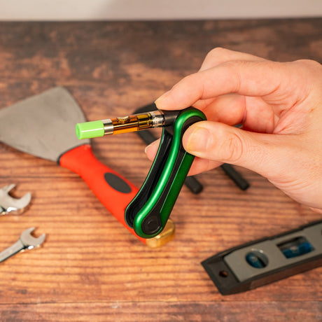 Hand holding a green Hinge, folding the cartridge back inside, on a wooden surface with tools in the background