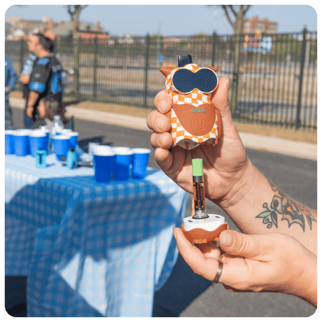 Person holding a Hoot owl vape device with a checkered pattern outdoors, near a table with blue cups.