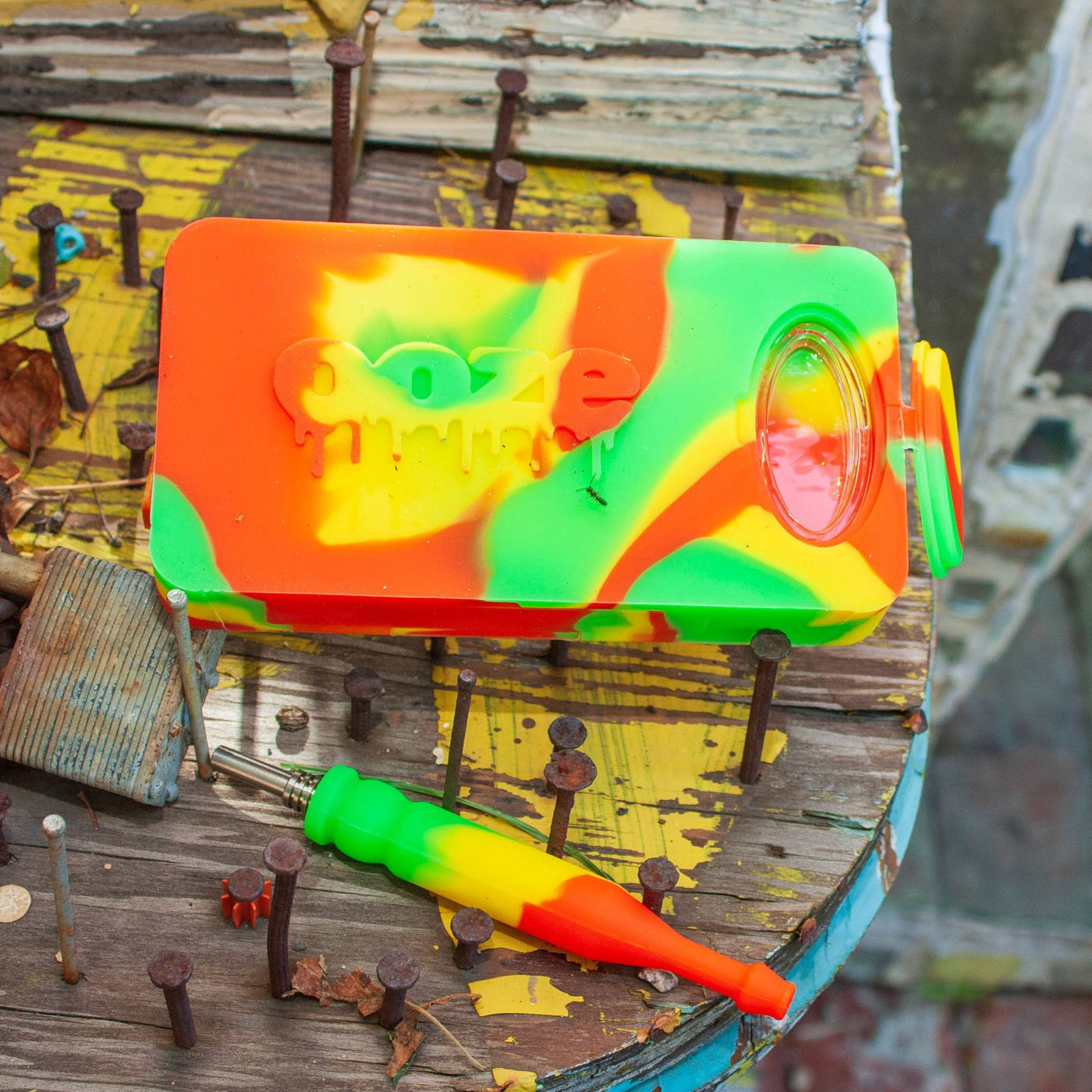 A rasta colored Ooze Slugger dab dugout is sitting on an outdoor table with rusty nails