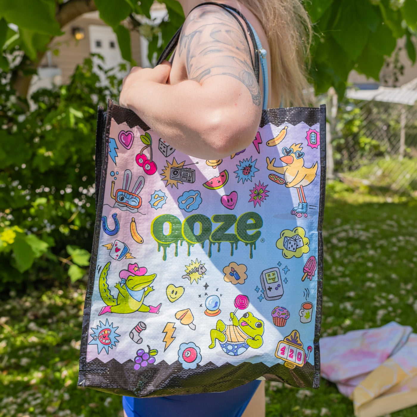 A person outdoors holding a reusable tote bag with a colorful cartoon print and the word 'ooze' in large green letters across the front.