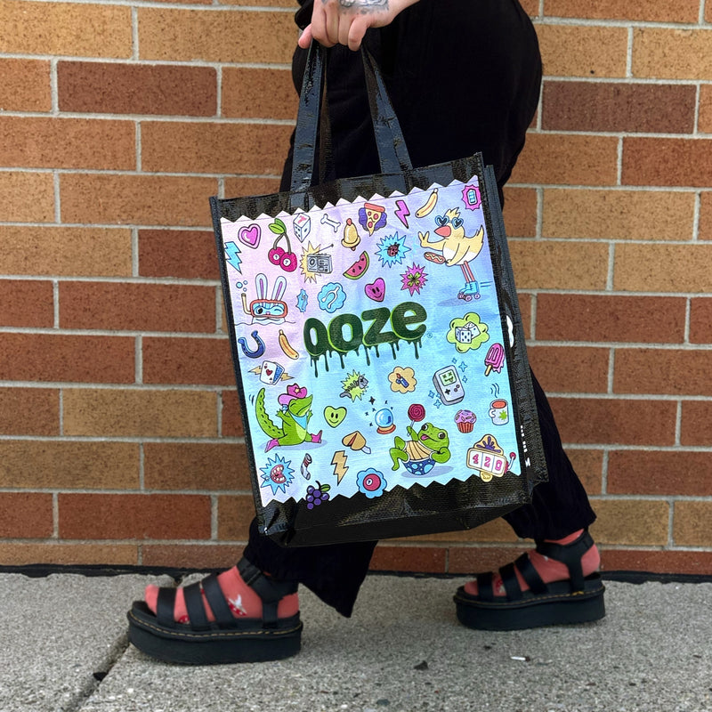 A person wearing black pants and pink-and-black platform sandals holds a black tote bag with a colorful cartoon-style graphic and the word Ooze printed on it, standing in front of a brick wall.