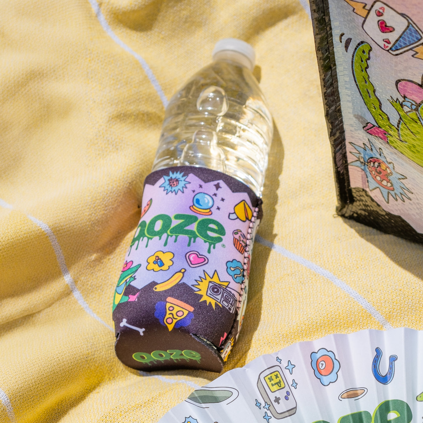 A water bottle in the Ooze Hotel Lucky slim can koozie is laying on a yellow blanket