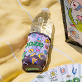 A water bottle in the Ooze Hotel Lucky slim can koozie is laying on a yellow blanket