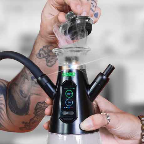 A hand holds the base of the black Ooze Ripley dab rig hookah, and a second hand pulls the carb cap off to reveal a plume of smoke