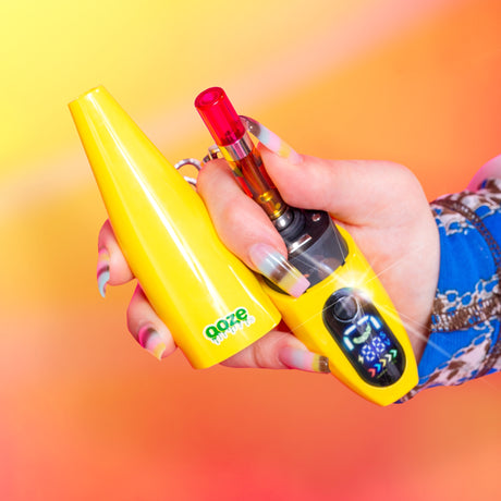 A hand holding the yellow cap and base of the Ooze Peel banana battery to show the cartridge inside, against a yellow orange gradient background