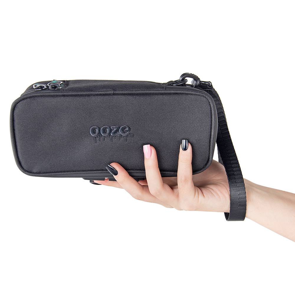 A girl with black and pink nails is holding a closed black Ooze travel pouch while wearing the wrist strap
