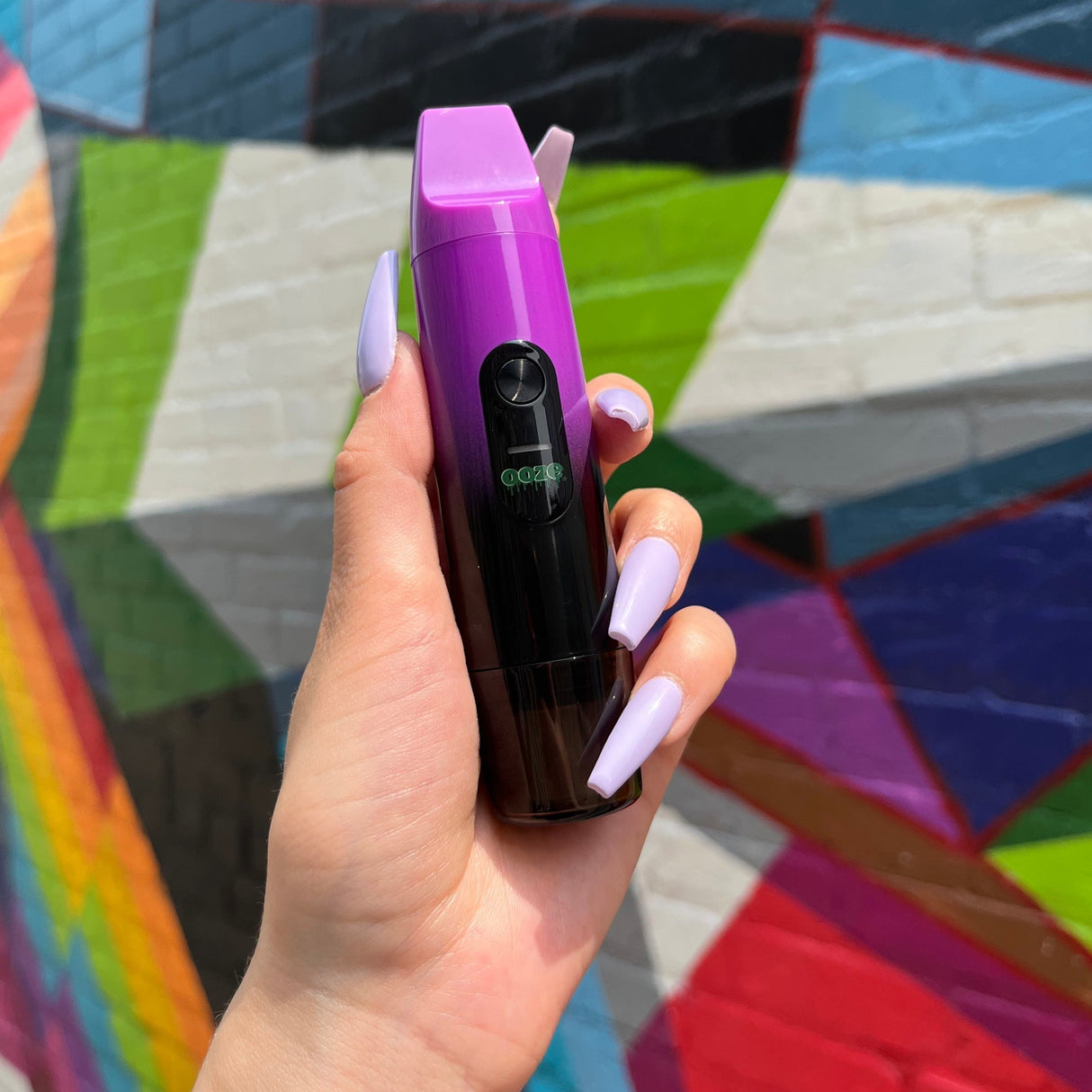 A girl's hand with long lilac acrylic nails holds the Galaxy Purple Ooze Booster in front of a colorful wall mural.