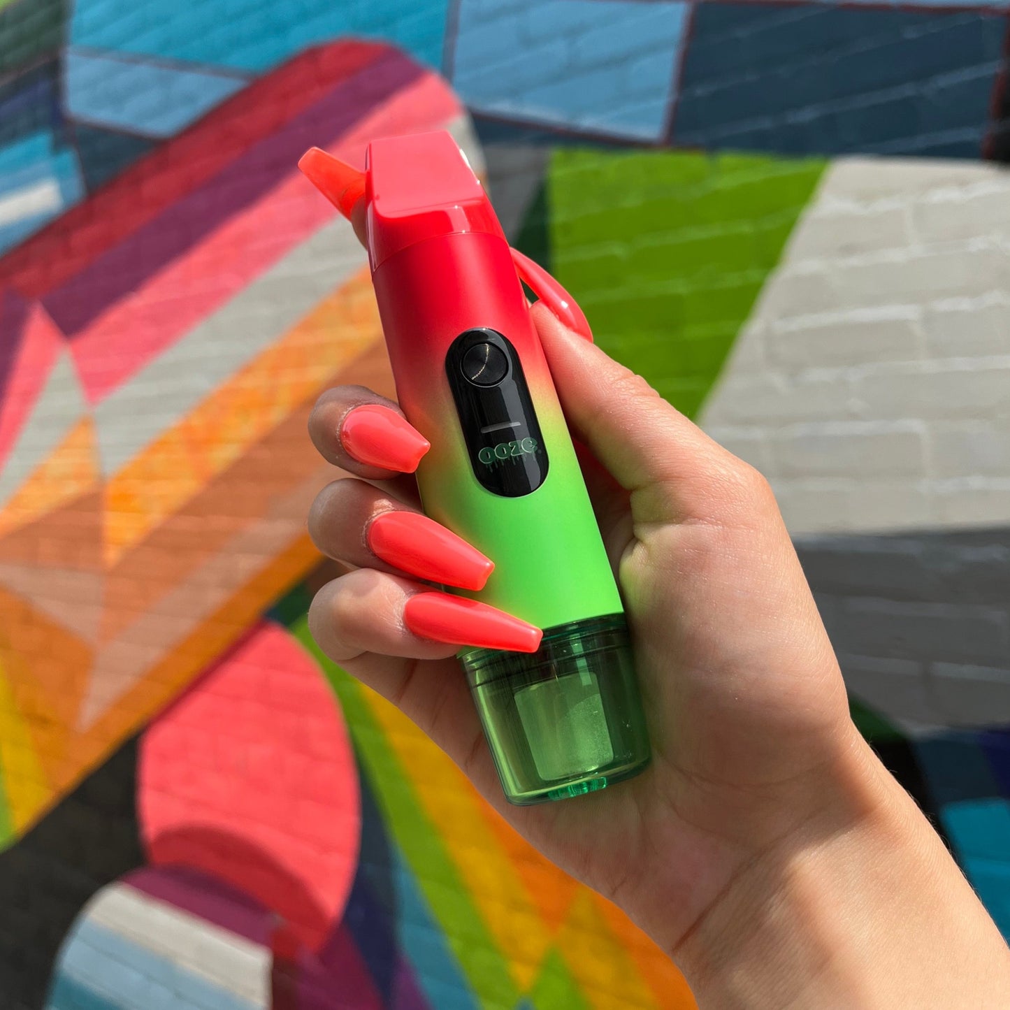 A girl's hand with long orange acrylic nails is holding the Rasta Ooze Booster in front of a colorful mural