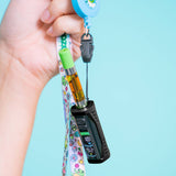 A hand holds a black Ooze Runt Mini vape device attached to a colorful patterned wrist strap and a blue retractable badge reel against a light blue background.