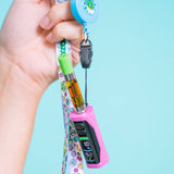 A hand holds a pink Ooze Runt Mini vape device attached to a colorful patterned wrist strap with a black clip and blue retractable badge reel, against a light blue background.