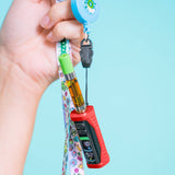 A hand holds a blue retractable vape lanyard attached to a red Ooze Runt Mini vape device with a green mouthpiece and a black clip, against a light blue background.