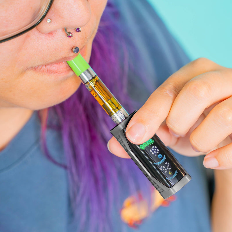 A close-up lifestyle shot of a person with purple hair and facial piercings holding a black Ooze Runt MIni vape pen to their lips. The vape pen is black with a bright green mouthpiece and a clear tank showing amber liquid, set against a solid light blue background.