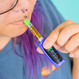 A close-up lifestyle shot of a person with purple hair and facial piercings holding a blue Ooze Runt Mini vape device to their lips. The vape has a bright green mouthpiece, a clear tank filled with amber-colored liquid, and a black digital display screen on the side.