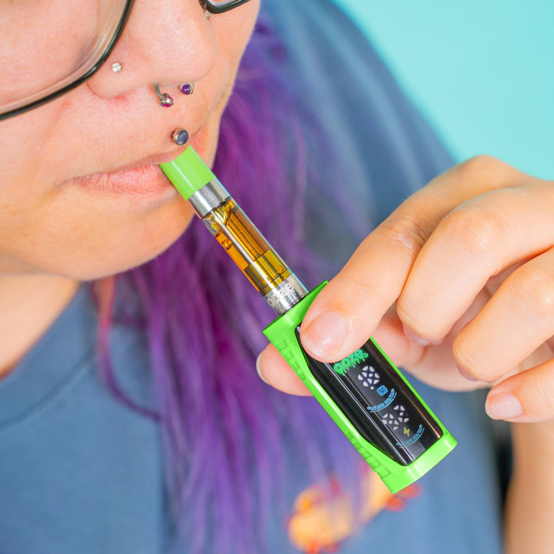 Close-up lifestyle shot of a person hitting an oil cartridge attached to a green Ooze Runt Mini vape battery with a digital screen