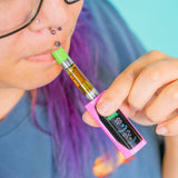 A close-up lifestyle shot of a person with purple hair and glasses vaping an oil cartridge on a pink Ooze Runt Mini battery. The vape device features a bright pink outer body, a black digital screen, and a green mouthpiece.