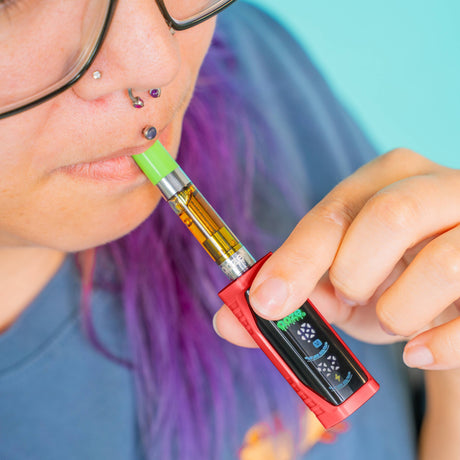 A close-up lifestyle shot of a person with purple hair and glasses using a red Ooze Runt Mini vape device with a green mouthpiece and a digital display screen.