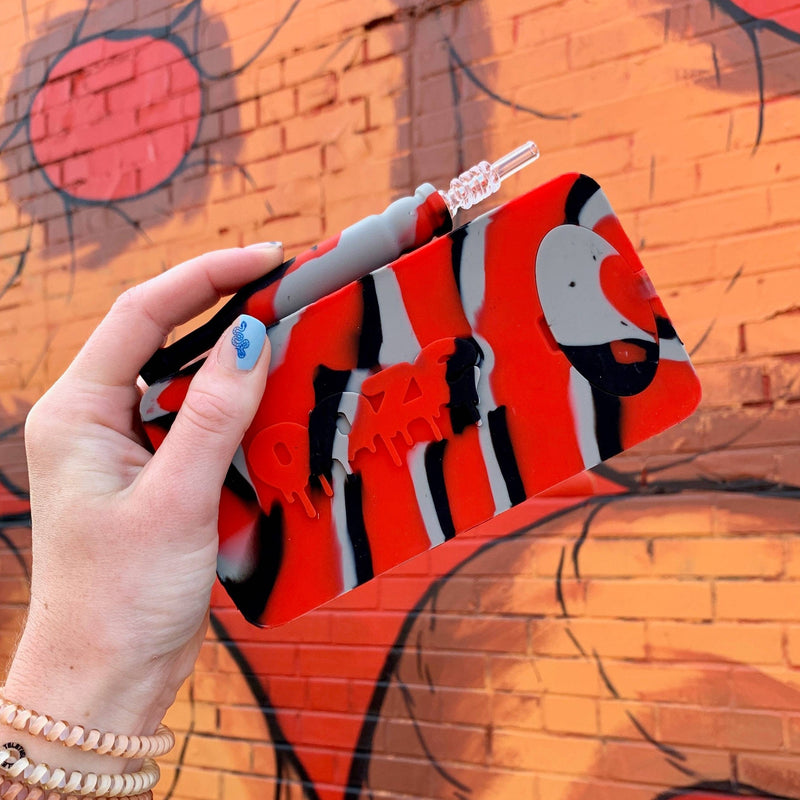 Hand holding a red, gray and black Ooze Slugger silicone dab dugout with the straw on top against a mural wall background