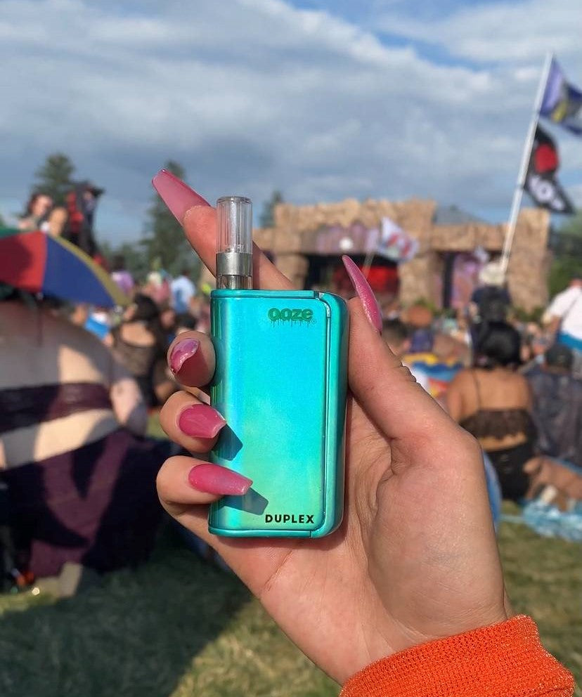 A close-up of a hand holding an arctic blue Ooze Duplex Pro outside at a music festival