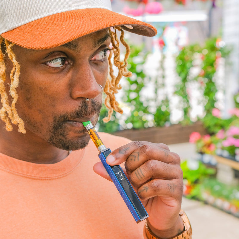 A close-up lifestyle shot of a man wearing a white and orange cap and an orange shirt, holding a blueOoze Quad 2 vape device to his mouth in an outdoor setting with blurred greenery and flowers in the background.