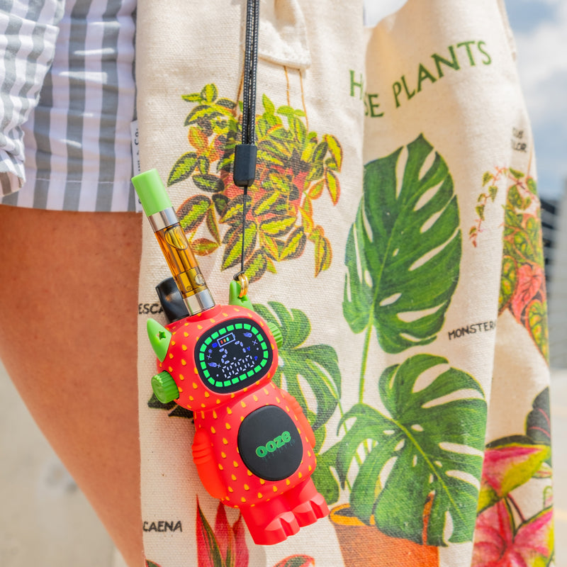 A strawberry ooze bot vape is hanging on a canvas tote bag with leaves printed on it