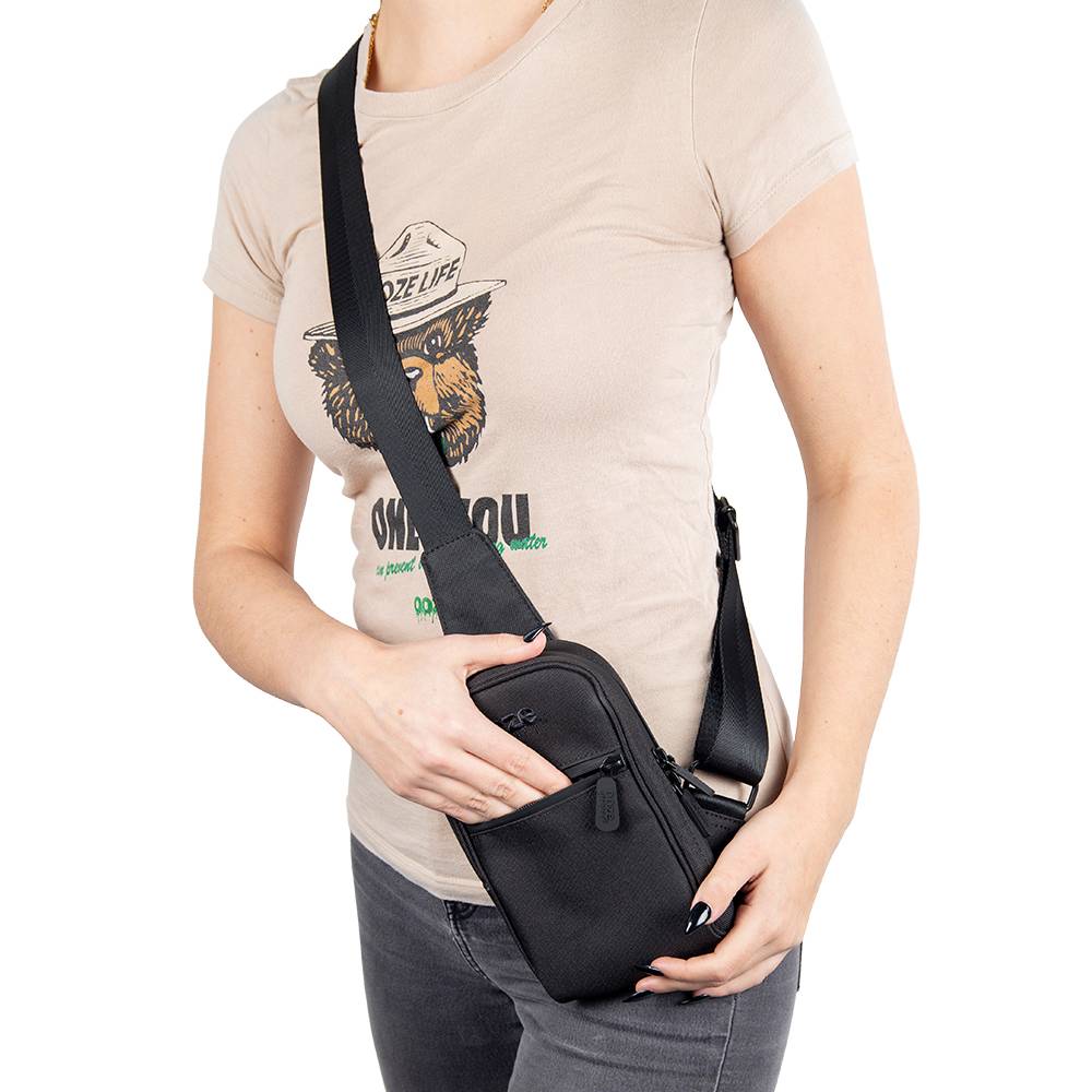 A girl is wearing a tan shirt and the black Ooze crossbody bag. She's sticking her hand into the front pocket