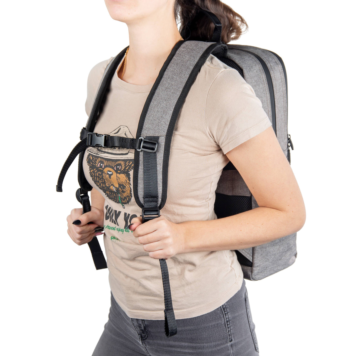 A girl in a tan shirt is wearing the gray Ooze backpack with the straps  buckled across her chest