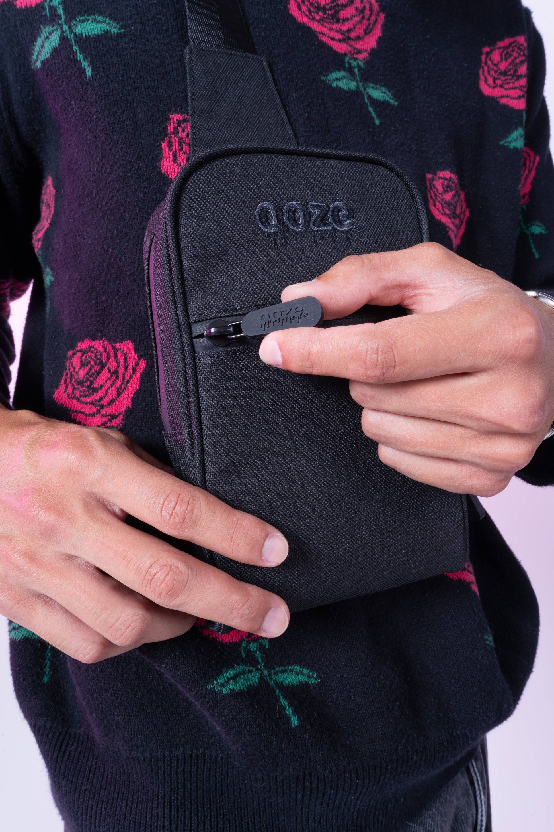 A guy in a rose sweater is wearing the black Ooze crossbody bag and unzipping the front pocket