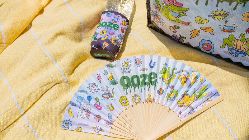 An expanded Ooze Hotel Lucky paper fan, slim can koozie on a water bottle, and reusable bag are laid on a yellow blanket