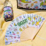 The Ooze Hotel Lucky paper fan is spread out, laying on a yellow blanket with a Hotel Lucky slim can koozie on a water bottle in the background