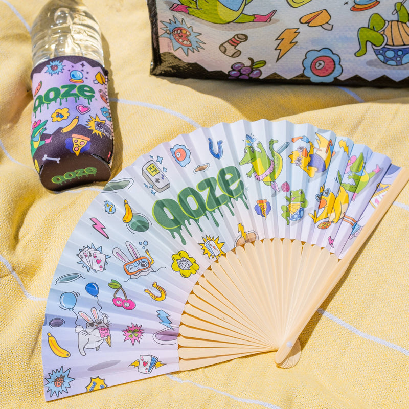 The Ooze Hotel Lucky paper fan is spread out, laying on a yellow blanket with a Hotel Lucky slim can koozie on a water bottle in the background