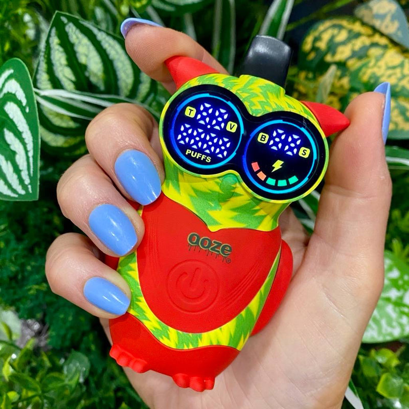 Inferno Ooze Hoot owl vape device held by a hand with blue nail polish against a green leafy background