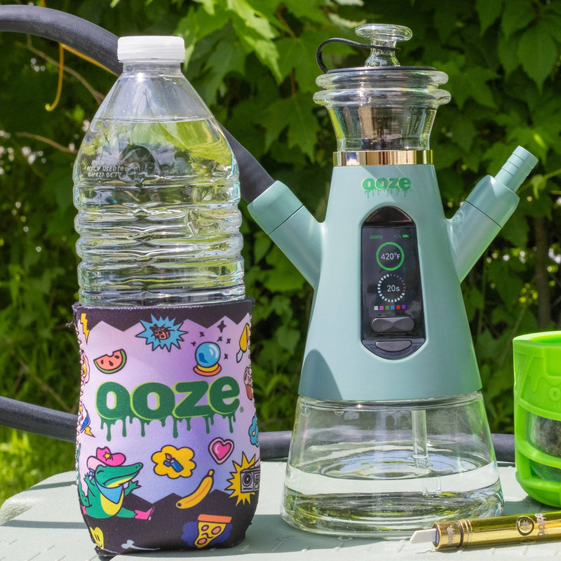 A mint green Ooze Vaporizer glass water pipe sits next to a clear plastic water bottle with a Hotel Lucky Ooze-branded sleeve, set outdoors on a table with green foliage in the background.