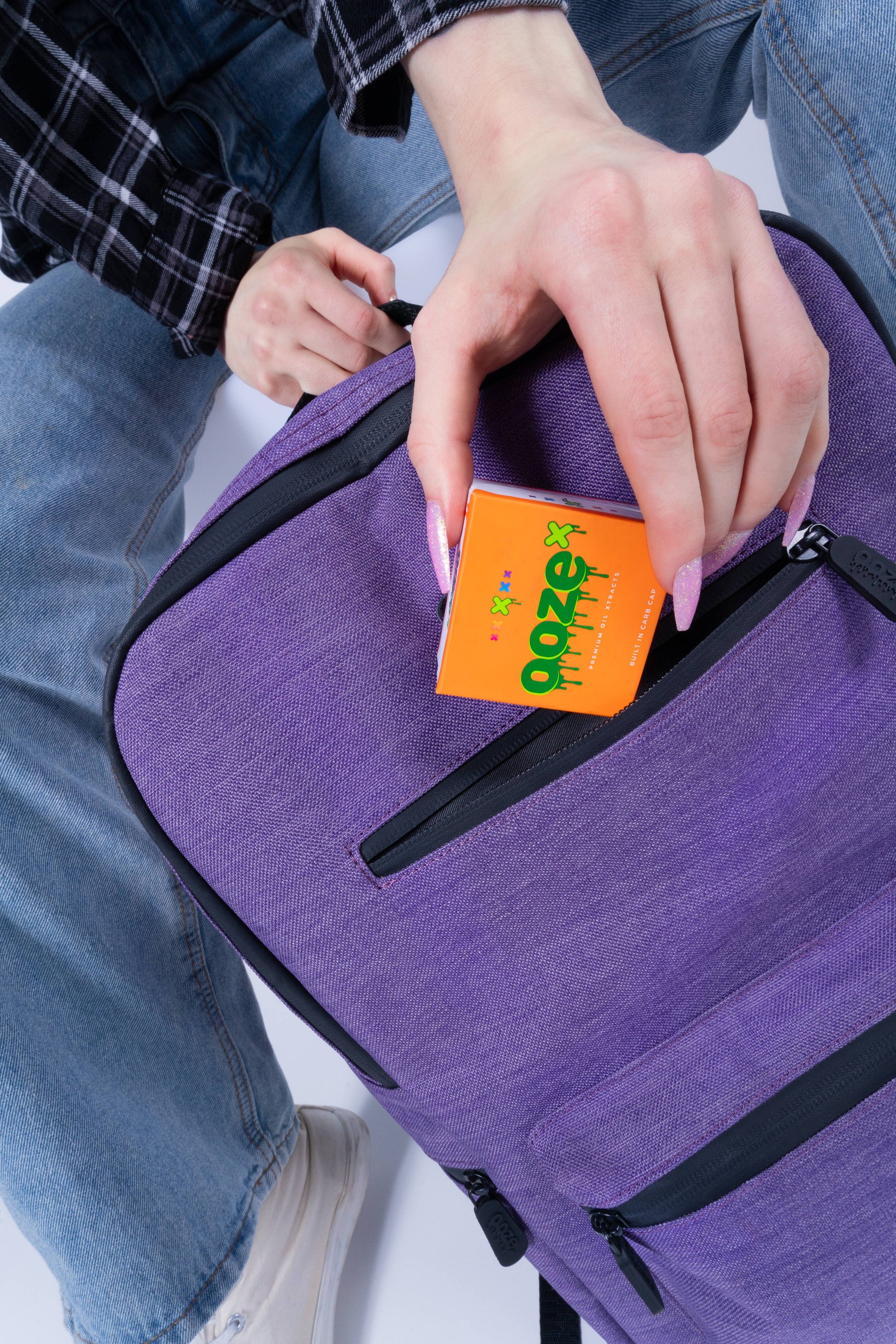 A girl is putting an OozeX box of concentrates into the top front pocket of the purple Ooze backpack
