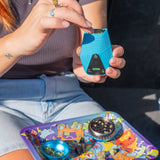 Person holding a blue Brink dry herb device with a colorful tray featuring a grinder on a blurred background
