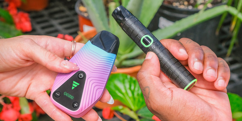 A close up of two hands holding a twilight Ooze brink and black Ooze pinch dry herb vapes in front of some flowers
